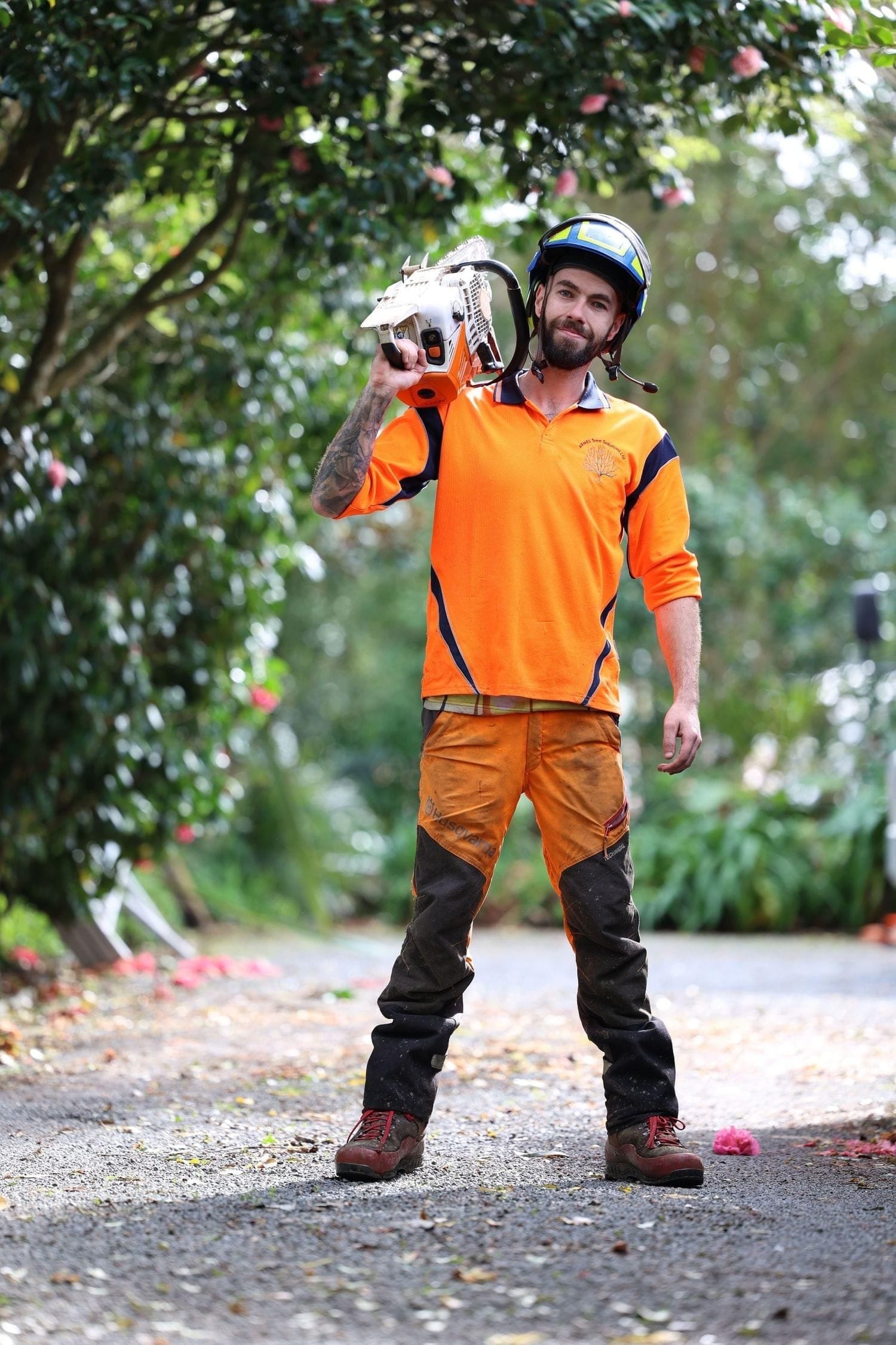 Arborist in orange work gear carrying a chainsaw on a tree-lined driveway