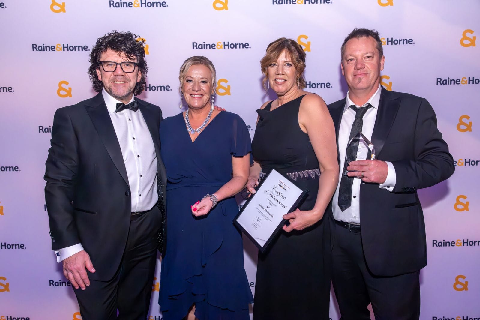 Four guests in black tie and evening dresses holding an award certificate at a media wall